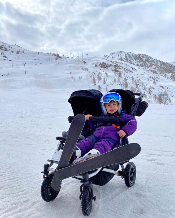 child dressed in ski suit with both skis on sitting in duet™ four wheeled double stroller at skiing field - Mountain Buggy life without limit